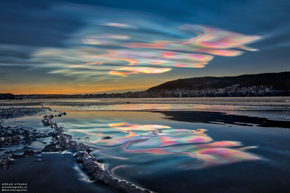 A landscape shows the sky above, shallow water in front,
and hills to the right. The sunset sky is filled with
beautifully multicoloured clouds. These clouds are clearly
reflected by the calm water in front. 
Please see the explanation for more detailed information.