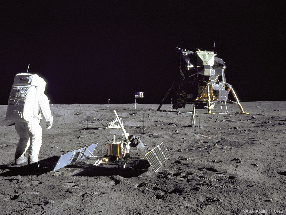 An astronaut is seen standing on the Moon next to 
an instrument with the lunar lander several metres in the 
distance. The dark of deep space covers the top of the frame. 
Please see the explanation for more detailed information.