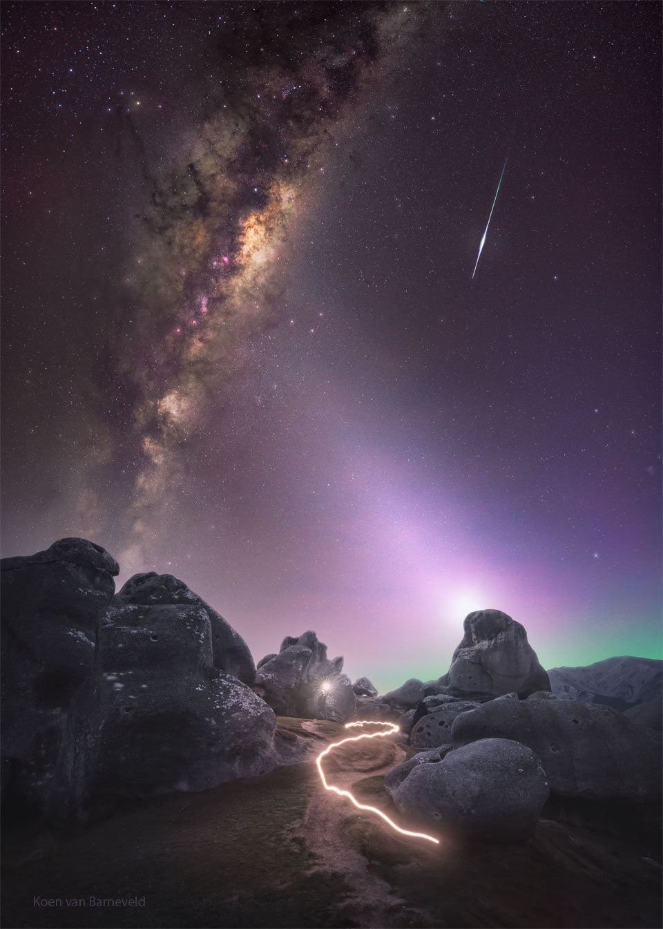 A skyscape is shown over a rocky landscape. In the starry sky
are the central band of our Milky Way Galaxy on the left, a 
meteor trail on the right, the dim band of zodiacal light 
in the centre, and the photographer holding a light just below
the centre. The path of the light is shown as a bright streak
in the bottom part of the frame. 
Please see the explanation for more detailed information.