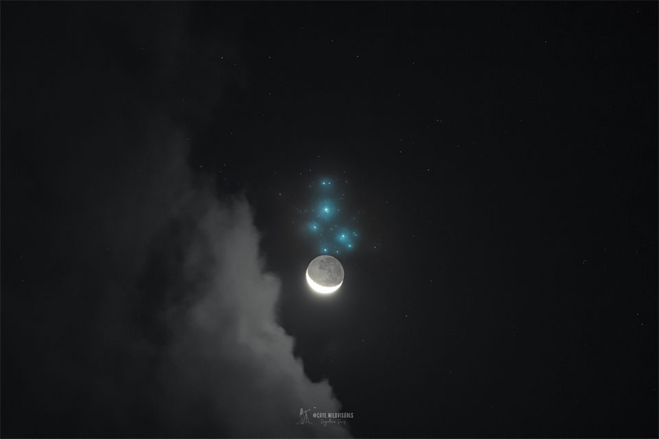 A dark night sky is shown with clouds on the left. 
Just above centre in blue is the Pleiades star cluster. 
Just below the Pleiades is a crescent moon, but bright 
enough so that you can see not only the brightly lit
crescent but, more faintly, the rest of the Moon.
Please see the explanation for more detailed information.
