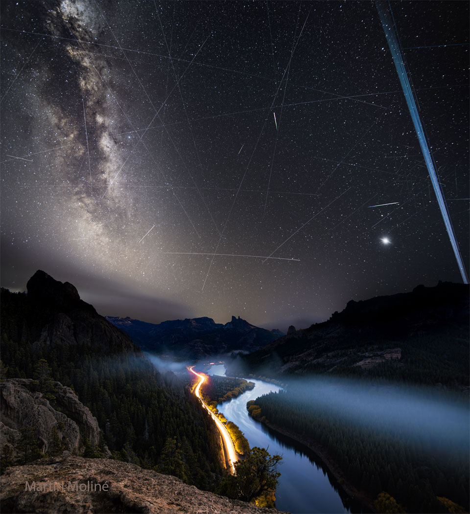 A night sky is shown with many stars and streaks.
In the foreground at the bottom are hills, a river, and
the red and white streaks of car lights.
Please see the explanation for more detailed information.