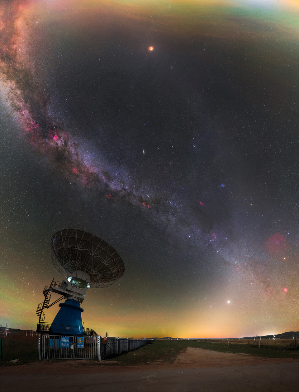 A wide angle image of the night sky is punctuated by
a radio telescope on the lower left. Two bands of light
run through the sky: the Milky Way and zodiacal light.
The bands intertwine like a double helix. Near the top
is an orange orb that is a totally eclipsed moon.
Please see the explanation for more detailed information.