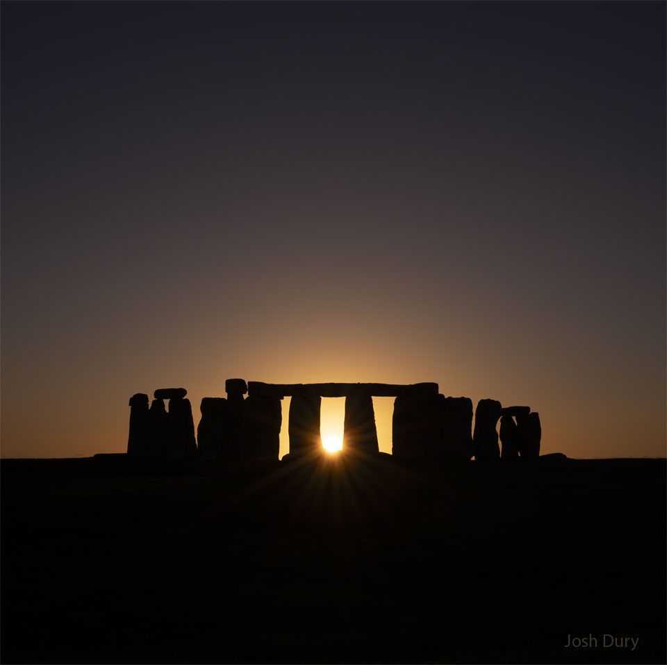 The sun is setting across a field in a clear sky.
In the field are the famous stones of Stonehenge. The
Sun peaks out from the centre of the stone array.
Please see the explanation for more detailed information.