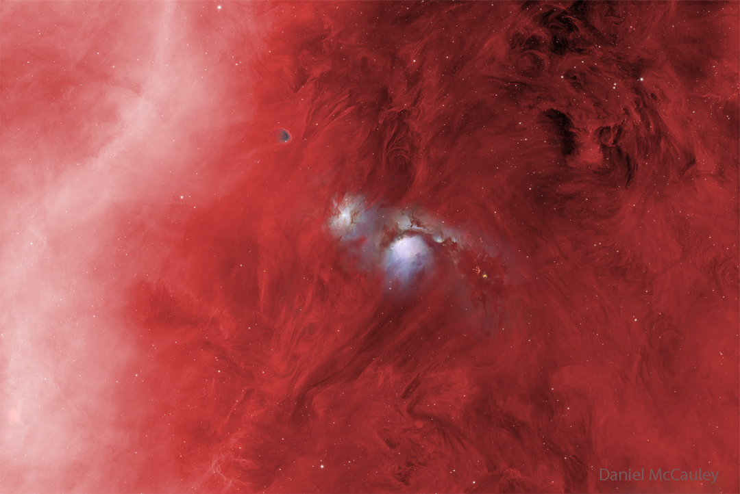 In a starfield glowing red, several features stand
out. On the right is a diffuse white band, and on the 
upper right is some dark dust. Most prominently, though,
are two blue structures in the centre, one of which has
a dark band running across near the middle. 
Please see the explanation for more detailed information.