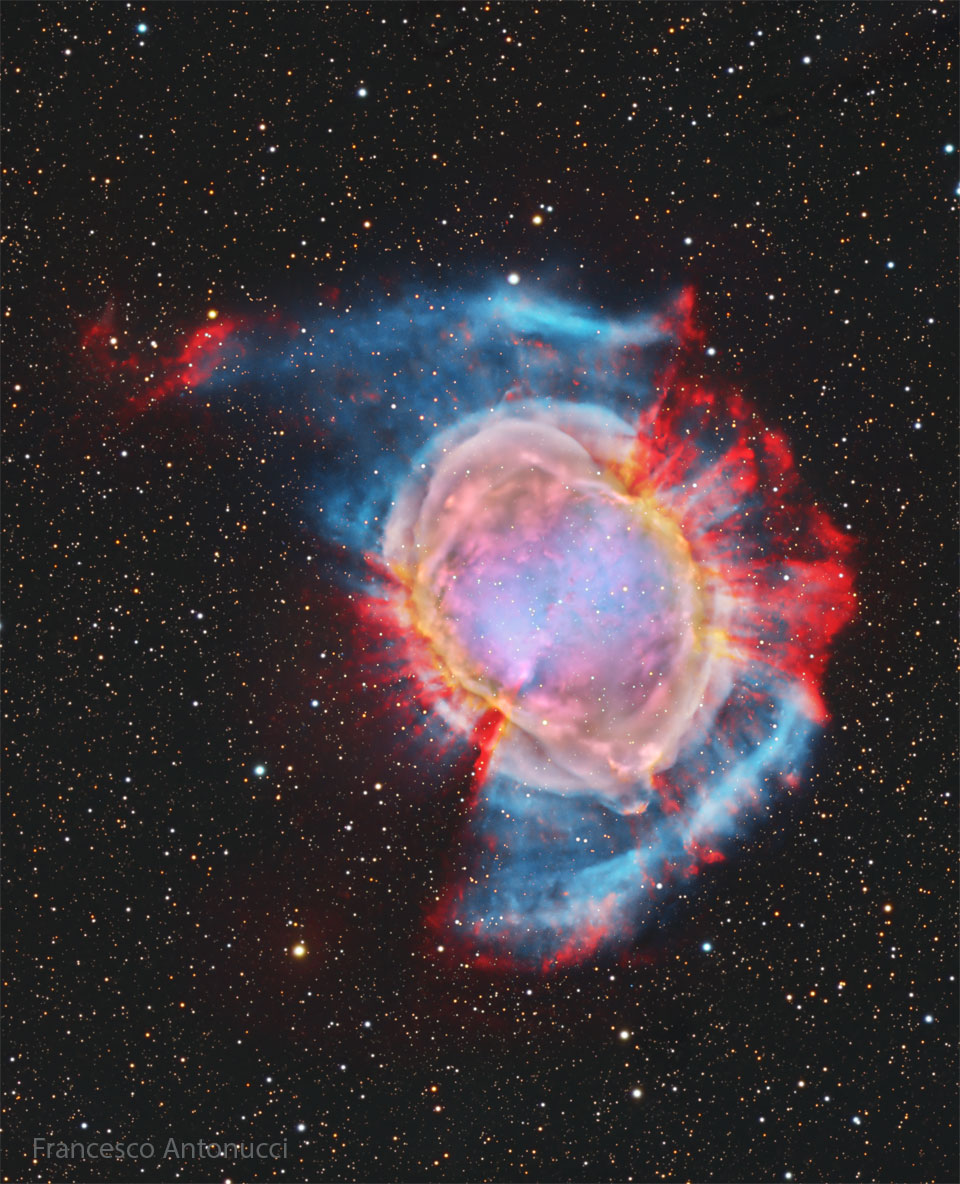 A starfield surrounds a large and colourful nebula.
Red spokes poke out from the outside of the nebula in
some directions, while a blue haze appears in other
directions. The nebula centre is multicoloured. 
Please see the explanation for more detailed information.