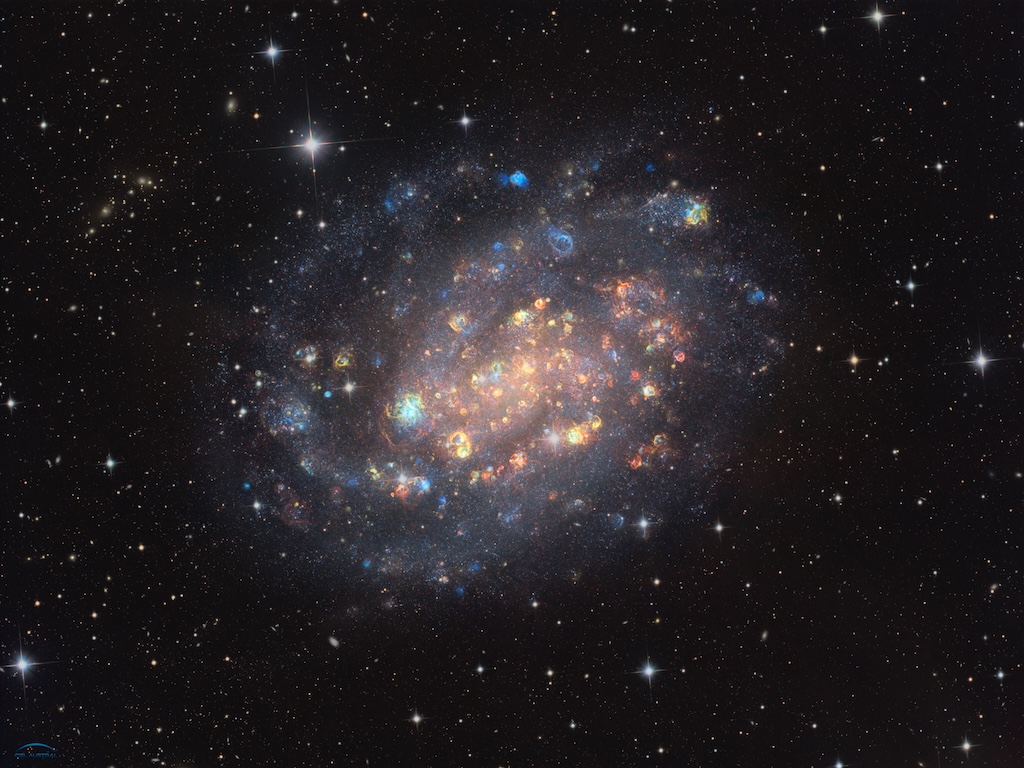 A bright spiral galaxy shows stars and colourful clous of gas
	  in red, green and blue colours. The background is a dark field of stars. 