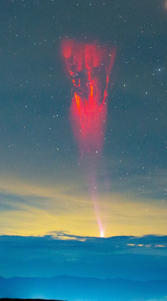 Lightning spreads like roots from a tree up into the atmosphere from the top of a cloud. Stars are scattered across the night sky above and the desert sits below the cloud deck.