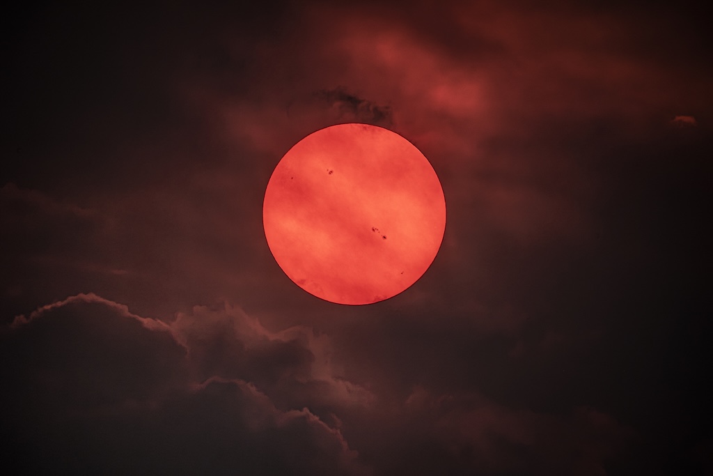 A deep red sun shows several sunspots.
	  Clouds and haze are visible. 