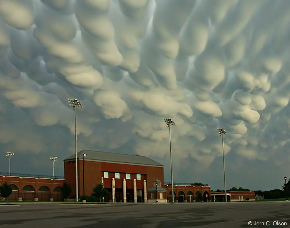 An unremarkable red building is seen past a large parking lot. 
Above them both are a bank of very unusual clouds with 
many nodules pointing down.
The scene is lit by sunlight from the side. 
Please see the explanation for more detailed information.