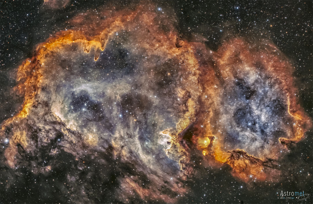 A red tinged nebula is shown in front of a starfield. 
Dust structures appear around the nebula's edge, and stars
are also seen near the nebula's centre.
Please see the explanation for more detailed information.