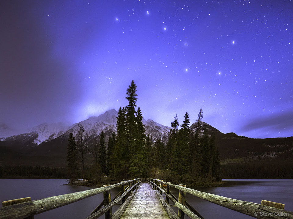 A landscape shows tall mountains in the distance and evergreen
trees nearby. Overhead is a star filled sky, with the stars of the 
Big Dipper easily apparent. A rollover image labels names for the 
Big Dipper stars.
Please see the explanation for more detailed information.