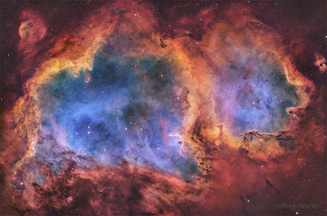 A starfield surrounds the edges of a large nebula.
The nebula, itself full of stars, has a blue glowing interior
and an orange periphery dotted with dust pillars. 
Please see the explanation for more detailed information.