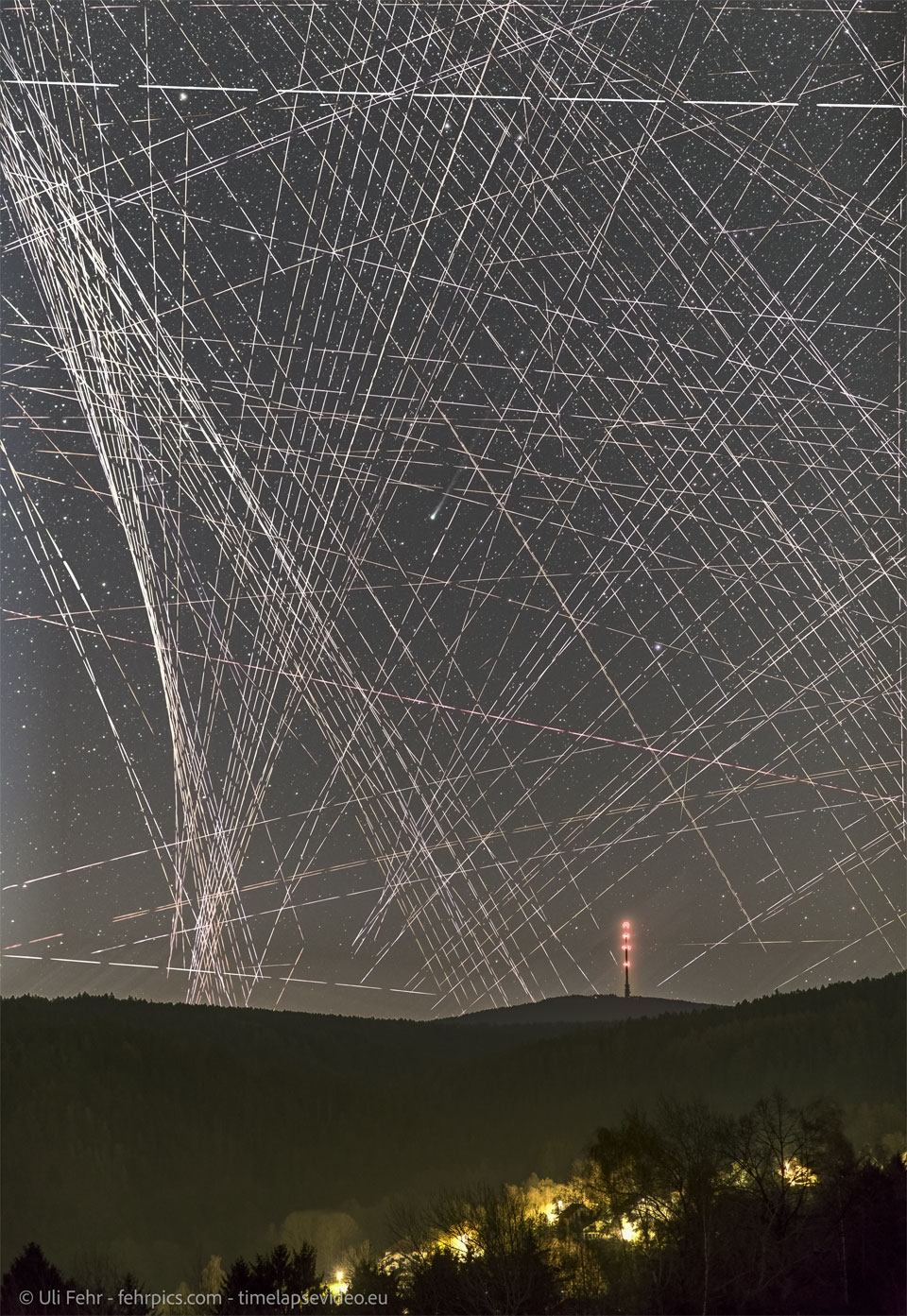 A night sky filled with lines and dashed-lines 
appears above a hilly landscape featuring a distant
lit-up tower. Near the centre of the image is a comet
shown by its small green coma and ion tail to the upper 
right.
Please see the explanation for more detailed information.