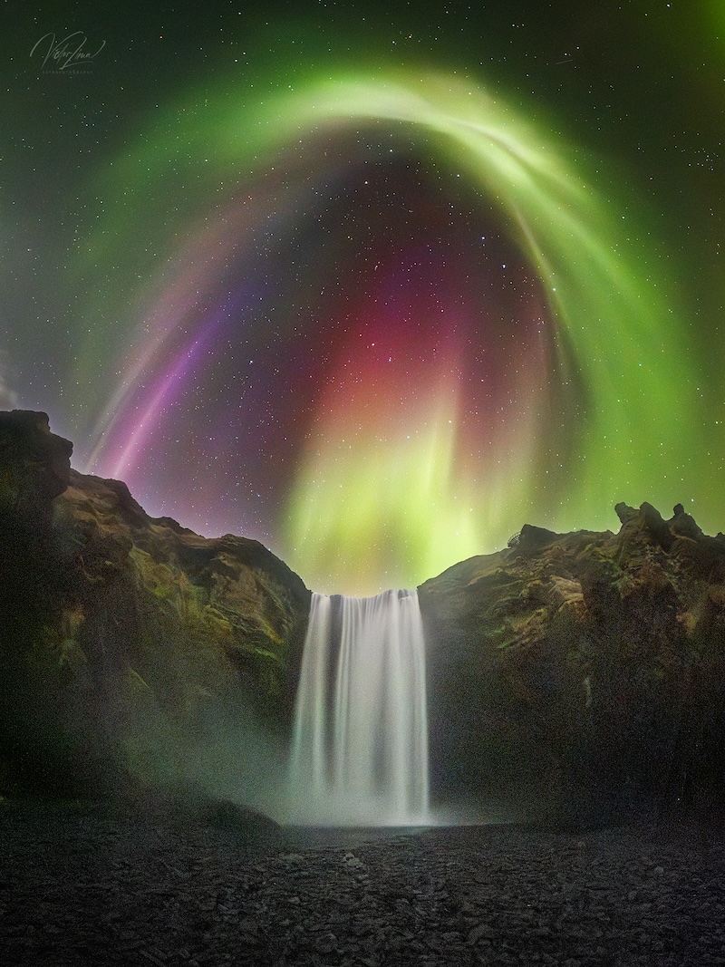 A night view of a waterfall with a colourful aurora in shades
	  of green, yellow, red and purple, and stars in the background.
