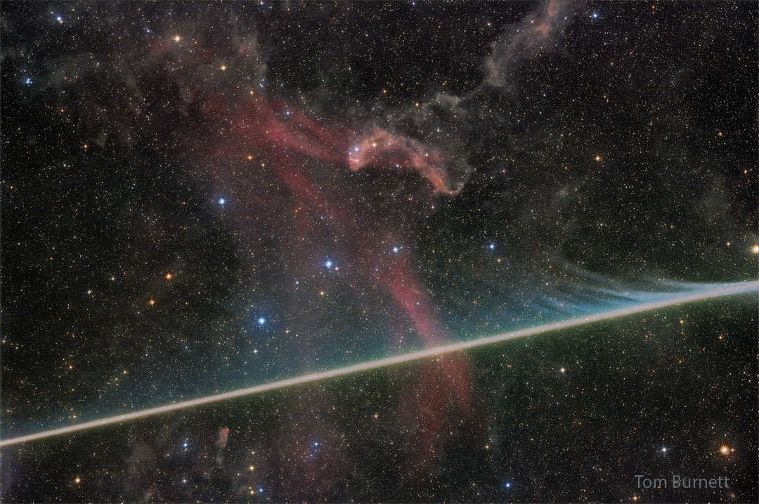 A dark starfield has a faint red nebula near 
the centre. A bright streak crosses the image 
horizontally from the lower left, filled with faint
evaporating wisps in many colours. 
Please see the explanation for more detailed information.