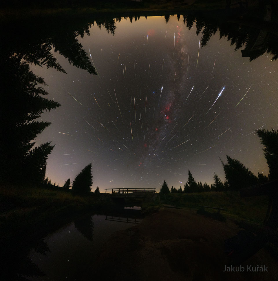 A starfield is bounded by trees on three sides and
a pond on the bottom. Amongst the stars are many streaks
of light appearing to emanate from a place on the sky
just above a foreground bridge. 
Please see the explanation for more detailed information.